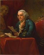 Benjamin Franklin sits reading, resting his elbows on a book covered table and with one hand under his chin. He wears a wig, glasses, and a blue coat.